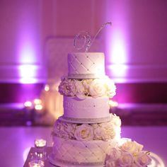 a three tiered wedding cake with white flowers on the table and purple lighting behind it