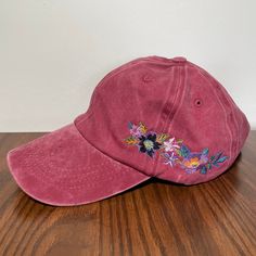 a pink baseball cap with embroidered flowers on the front and side, sitting on a wooden surface