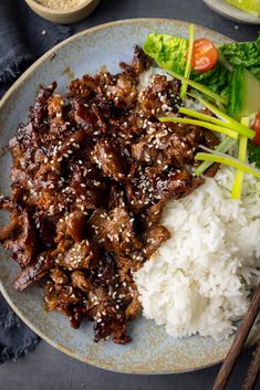 a plate topped with meat and rice next to chopsticks on a blue table cloth