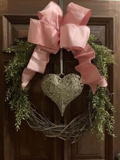 a wreath with a heart hanging from it's side on a wooden door decorated with greenery and pink ribbon