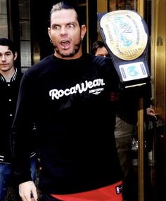 a man holding up a wrestling belt in front of a doorway with other men behind him