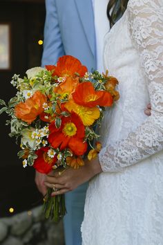 a bride holding a bouquet of orange and yellow flowers next to her groom in a blue suit