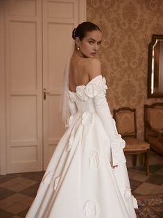 a woman in a white wedding dress standing next to a chair and looking off into the distance