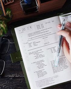 a person holding a pen in their hand while writing on an open notebook next to some glasses