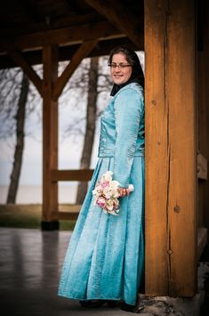 a woman in a long blue dress standing next to a wooden structure and holding a bouquet