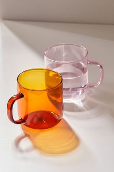 two glass mugs sitting next to each other on a white counter top, one has a red handle and the other is yellow