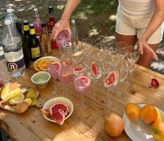 a woman is pouring wine into glasses on a wooden table with fruit and other drinks