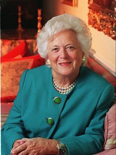 an older woman sitting on a couch wearing a green coat and pearls necklaces, smiling at the camera