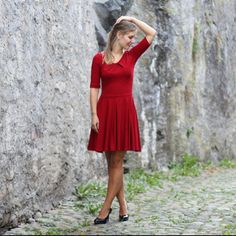 a woman in a red dress standing next to a stone wall with her hand on her head