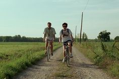 two men riding bikes down a dirt road next to a lush green field and telephone poles