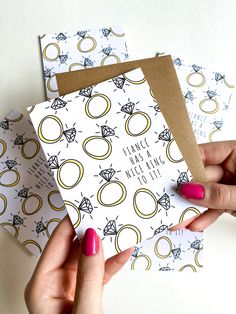 two hands holding envelopes that have wedding rings on them, one with pink nail polish and the other with gold