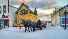 two horses pulling a carriage down a snow covered street in front of colorful houses with christmas wreaths