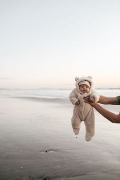 a man holding a baby wearing a teddy bear hat and scarf on the beach with water in the background