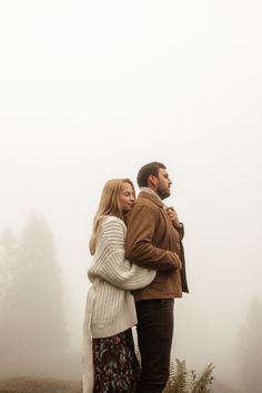 a man and woman standing on top of a hill with fog in the air behind them