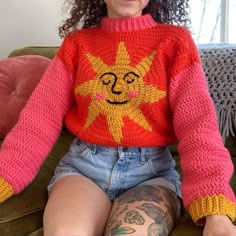 a woman sitting on top of a green couch wearing a red sweater with a yellow sun drawn on it