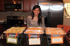 a woman standing in front of four trays of food on top of a kitchen counter