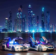 two cars parked next to each other in front of a city skyline at night with lights on