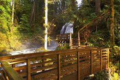 a wooden bridge over a river in the woods with waterfall behind it and stairs leading up to them