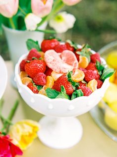 a white bowl filled with fruit sitting on top of a table next to some flowers