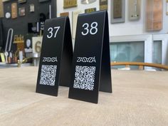 two black signs sitting on top of a wooden table next to each other with qr code printed on them