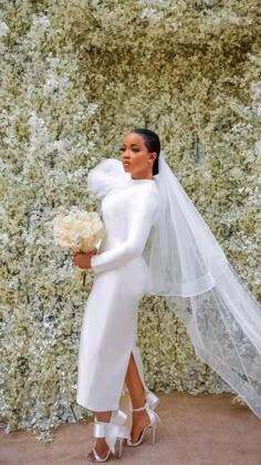 a woman in a white wedding dress holding a bouquet and standing next to a green wall