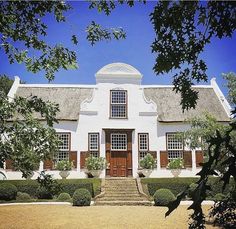 a large white house with brown shutters on the front and side doors, surrounded by hedges