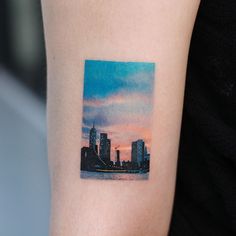 a city skyline tattoo on the left inner arm and upper arm, with an orange sunset in the background