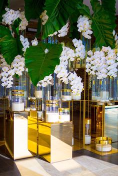 white flowers and candles are in glass vases with gold bases on the floor, along with greenery