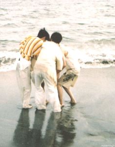 three people standing on the beach with their arms around each other and looking at the water