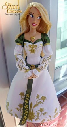 the doll is wearing a white dress with gold trimmings and a green ribbon