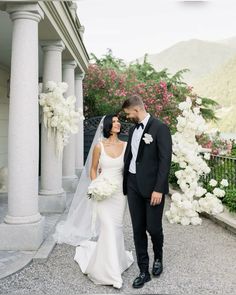 a bride and groom standing in front of some white flowers on the side of a building