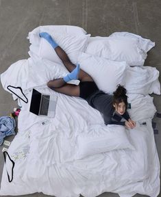 a woman laying on top of a white bed covered in pillows and blankets next to a laptop computer