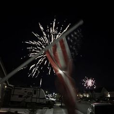 an american flag is waving in the wind with fireworks behind it and cars parked on the side of the road