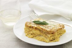 a white plate topped with lasagna covered in cheese and sage leaves next to a glass of water