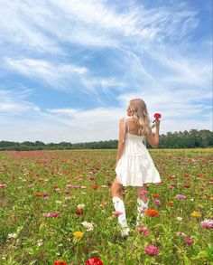 a girl in a field with flowers holding a flower up to her face and looking at the sky