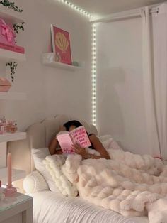 a woman laying in bed reading a pink book with lights on the wall behind her