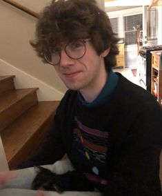 a man with glasses holding a cat in his lap while sitting on the floor next to stairs