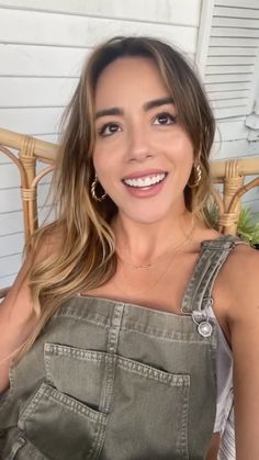 a smiling woman in overalls sitting on a chair with her hands behind her back