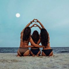 three women in bikinis sitting on the beach making a heart shape with their hands