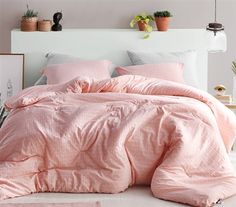 a bed with pink comforter and pillows in a white room next to potted plants