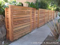 a wooden fence with plants growing on top of it next to a sidewalk and trees