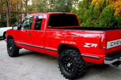 a red pick up truck parked in a parking lot next to some trees and bushes