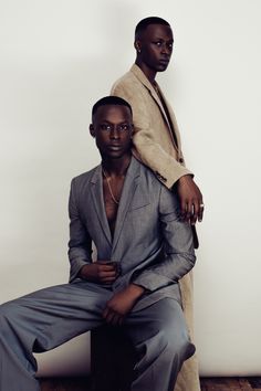 two men in suits sitting next to each other on a wooden floor and one is wearing a tan coat