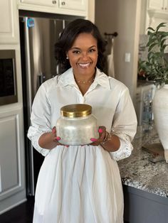 a woman in a white dress is holding a gold jar and smiling at the camera