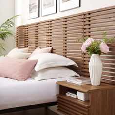 a white vase with pink flowers sitting on top of a bed next to a wooden headboard