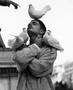 black and white photograph of a woman holding birds on her head while pigeons fly overhead