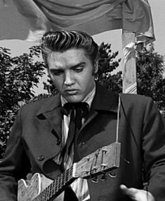 a man in a suit and tie holding a guitar near a flag with his hand