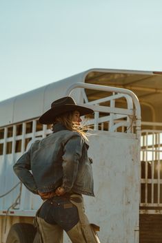 a woman wearing a cowboy hat and jeans is standing in front of a horse trailer