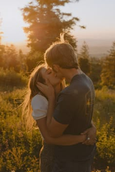 a young man and woman kissing in the grass with trees in the background at sunset