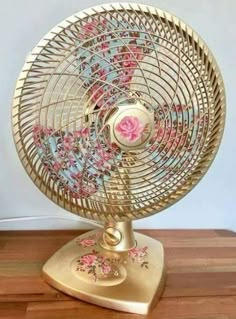 a gold fan with pink flowers on it sitting on top of a wooden table next to a wall